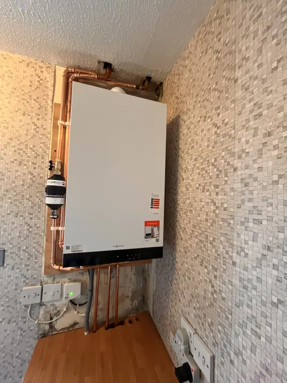 A wall-mounted combi boiler with a magnetic system filter and copper pipework running down into the floor.