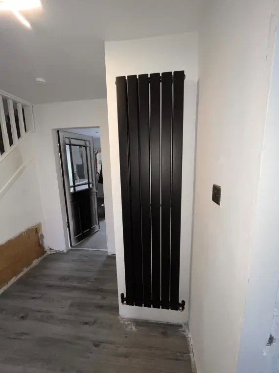A tall matte black vertical column radiator fitted in a hallway alcove beside a staircase.