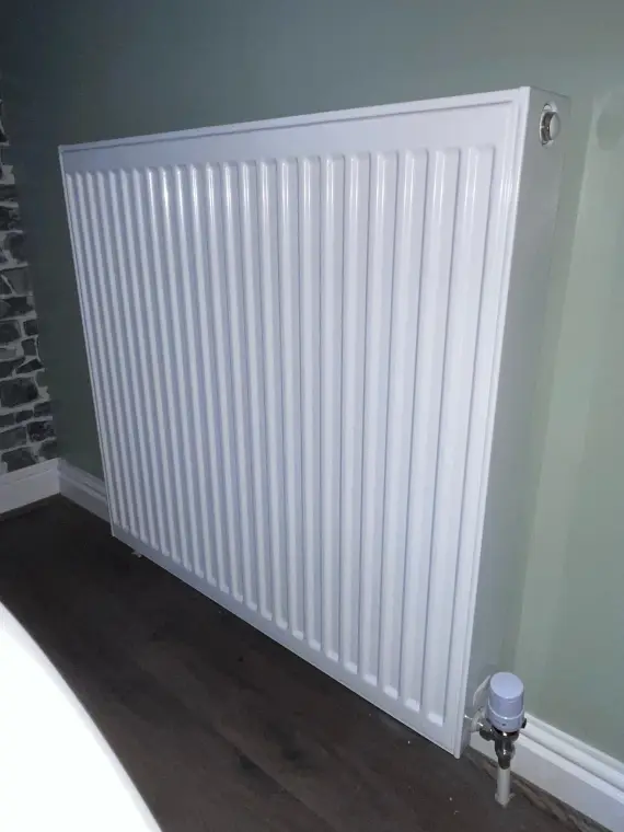 A white horizontal panel radiator on a sage green wall, with an exposed stone section to its left.